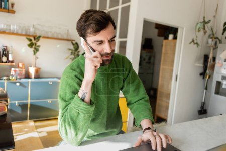 Bärtiger und tätowierter Freiberufler im Pullover spricht auf dem Smartphone, während er zu Hause am Laptop arbeitet 