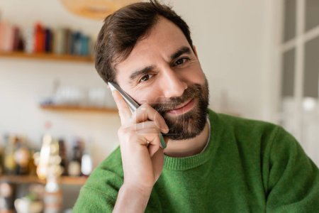 Porträt eines fröhlichen brünetten und bärtigen Mannes im grünen Pullover, der zu Hause mit dem Smartphone spricht 