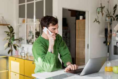 Bearded and tattooed freelancer in casual jumper talking on smartphone and using laptop at home
