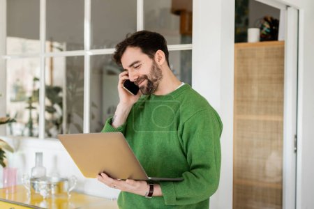 Positive brunette and bearded freelancer in jumper talking on smartphone and holding laptop at home 