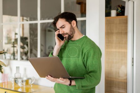 Focused freelancer in green jumper holding laptop and talking on smartphone at home 