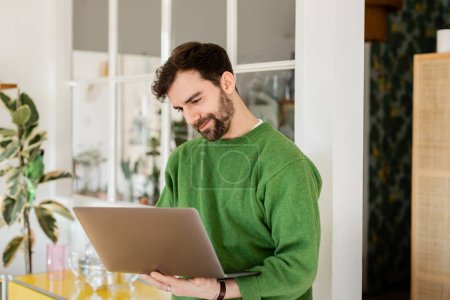 Positive brunette and bearded entrepreneur in green jumper looking at laptop while working at home