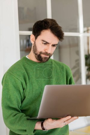 Portrait of focused and bearded freelancer in casual green jumper looking at laptop, remote work