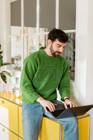 bearded freelancer in green jumper and jeans using laptop while working at home, remote work