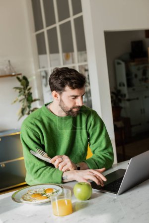 Photo for Bearded and tattooed freelancer in jumper holding cutlery and using laptop at home - Royalty Free Image