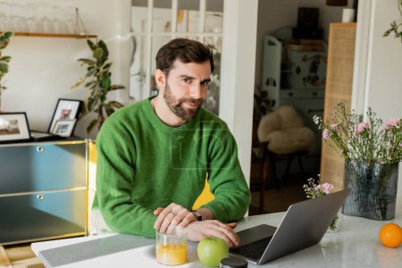 Bärtiger Mann im Pullover blickt in die Kamera, während er Laptop in der Nähe von Orangensaft in der modernen Küche benutzt
