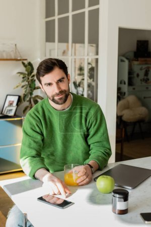 Brünette und bärtige Mann in grünem Pullover hält ein Glas Orangensaft und arbeitet zu Hause 