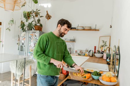 Téléchargez les photos : Heureux homme barbu en pull et jeans couper du poivre frais tout en cuisinant le petit déjeuner dans la cuisine - en image libre de droit