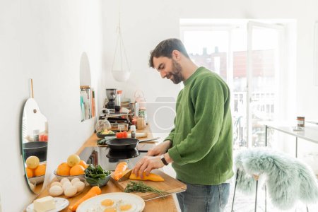 Foto de Sonriente hombre en jersey verde corte pimienta fresca mientras se cocina el desayuno en la cocina moderna - Imagen libre de derechos
