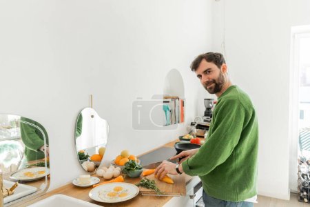 Foto de Hombre feliz en jersey verde cortar pimienta fresca mientras se cocina el desayuno en la cocina moderna - Imagen libre de derechos