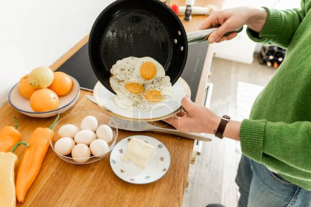 Foto de Vista recortada del hombre sosteniendo la sartén con huevos fritos cerca del plato mientras cocina el desayuno por la mañana - Imagen libre de derechos