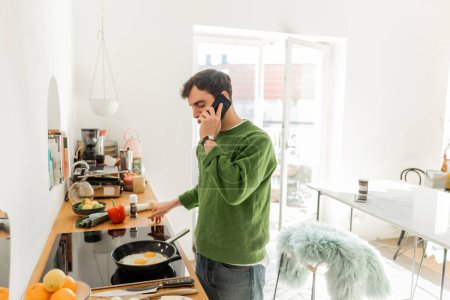 Foto de Morena hombre en jersey y jeans hablando en el teléfono inteligente mientras se cocina el desayuno en la cocina en casa - Imagen libre de derechos