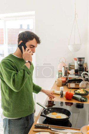 Foto de Hombre barbudo en jersey y jeans hablando en el teléfono inteligente mientras se cocina el desayuno en la cocina en casa - Imagen libre de derechos