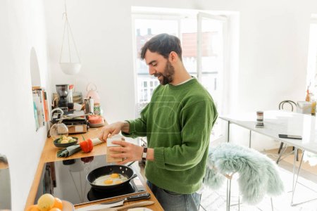 Foto de Barbudo hombre en verde jersey condimento huevos fritos mientras cocina el desayuno por la mañana - Imagen libre de derechos