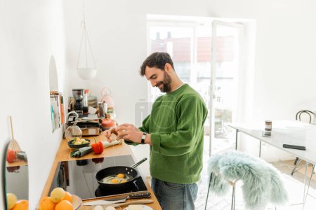 Foto de Hombre barbudo alegre en ropa casual vertiendo huevo en la sartén mientras cocina el desayuno - Imagen libre de derechos