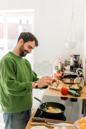 Foto de Hombre barbudo alegre vertiendo huevo en la sartén mientras se cocina cerca de alimentos frescos desenfocados en la encimera - Imagen libre de derechos
