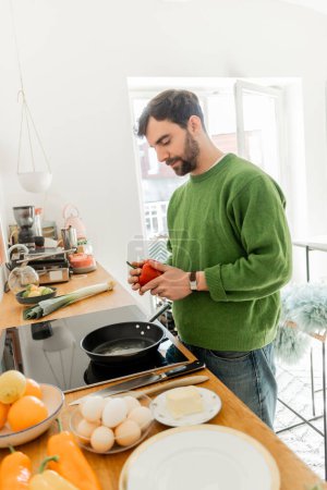 Bearded man in jumper holding fresh bell pepper near frying pan, blurred eggs and butter on worktop