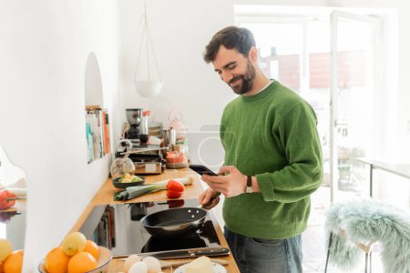 Positive and bearded man in casual clothes using smartphone while cooking breakfast 