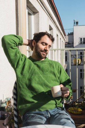 Relaxed bearded man with closed eyes in green pullover and jeans holding cup of coffee in morning