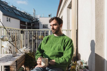 Entspannter Mann in grünem Pullover und Jeans hält morgens eine Tasse Kaffee auf dem Balkon