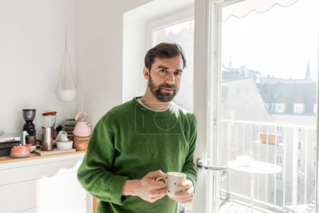 Porträt eines bärtigen Mannes im Pullover, der eine Tasse Kaffee hält und in der modernen Küche in die Kamera blickt