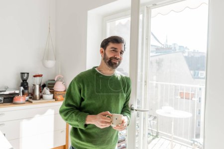 Fröhlicher bärtiger Mann im Pullover mit Tasse Kaffee und Blick in die Kamera 