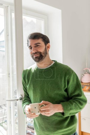 Positive bearded and brunette man in green jumper holding cup of coffee while standing near window