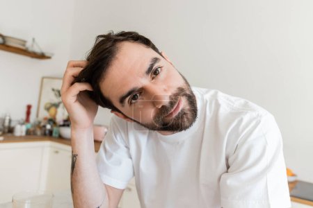 Portrait of bearded and tattooed man in white t-shirt touching hair and looking at camera 