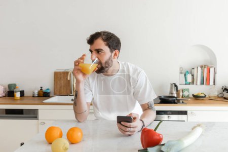 Bärtiger und tätowierter Mann in weißem T-Shirt trinkt frischen Orangensaft und benutzt Smartphone 