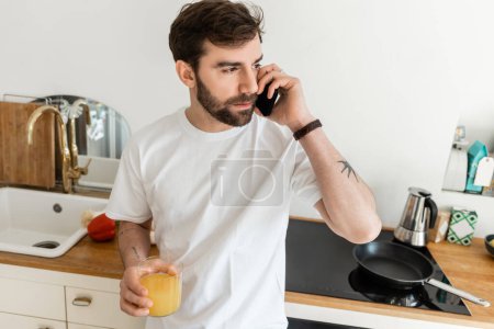 Tätowierter bärtiger Mann im weißen T-Shirt spricht mit Smartphone und hält ein Glas frischen Orangensaft in der Hand 