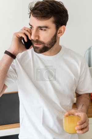 Porträt eines tätowierten Mannes im weißen T-Shirt, der mit dem Smartphone spricht und ein Glas Orangensaft in der Hand hält