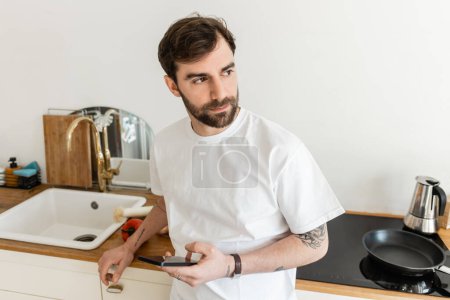 Foto de Hombre barbudo y tatuado en camiseta blanca mirando hacia otro lado y sosteniendo el teléfono inteligente - Imagen libre de derechos