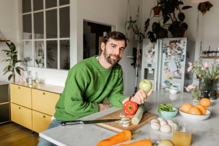 Bearded man in green jumper looking at camera while holding apple in modern kitchen at home 