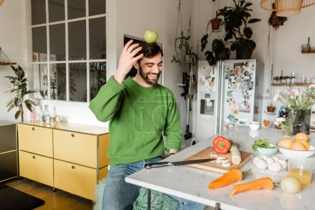 Fröhlicher und bärtiger Mann im grünen Pullover balanciert mit Apfel auf dem Kopf in moderner Küche 