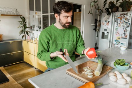 Bearded tattooed man in green jumper holding knife and bell pepper while cooking in modern kitchen