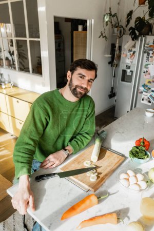 Positiver Mann im Pullover blickt in die Kamera, während er zu Hause in der modernen Küche neben frischen Lebensmitteln sitzt 