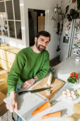 Positive man in jumper looking at camera while sitting near fresh food in modern kitchen at home  puzzle #657147948