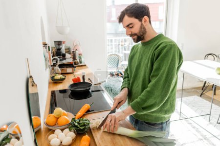 Photo for Young bearded man in casual clothes cutting leek near eggs and fresh food on worktop at home - Royalty Free Image