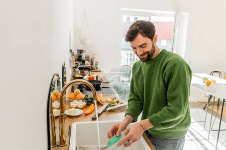 Cheerful and tattooed bearded man in green jumper and jeans washing plate at home 