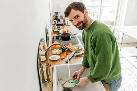 Lächelnder bärtiger Mann in lässiger Kleidung schaut in die Kamera, während er zu Hause Teller wäscht 
