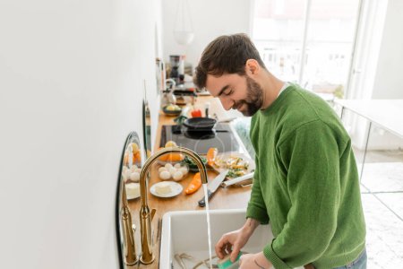 Lächelnder und bärtiger Mann in grünem Pullover wäscht zu Hause Geschirr 