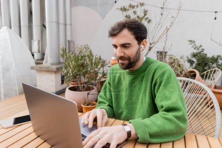 freelancer in green jumper using earphone and laptop while working on rooftop of house terrace 