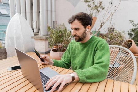 Freelance barbu en pull vert décontracté à l'aide d'écouteurs sans fil, smartphones et ordinateur portable 