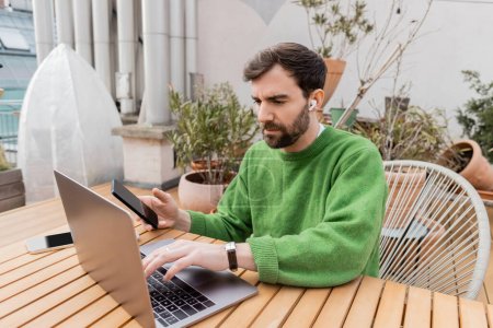 Entrepreneur barbu dans écouteur et pull vert tenant smartphone et en utilisant un ordinateur portable en terrasse de la maison 