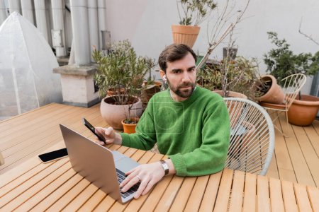 Téléchargez les photos : Bearded freelancer in wireless earphone in green jumper using smartphone and working on laptop - en image libre de droit