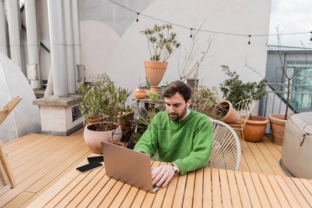 Téléchargez les photos : Entrepreneur en écouteur sans fil à l'aide d'un ordinateur portable tout en travaillant près des smartphones sur la terrasse de la maison - en image libre de droit