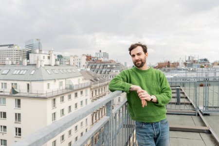 schöner Mann in grünem Pullover schaut weg, während er auf der Dachterrasse in Wien steht