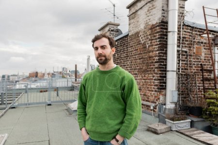 Bearded man standing on rooftop terrace of house with blurred cityscape at background in Vienna