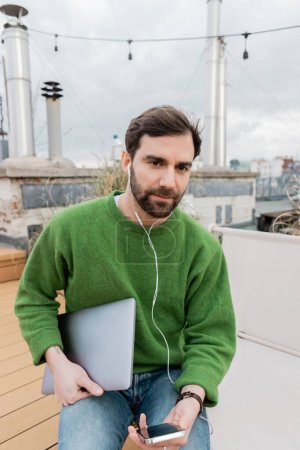 freelancer in earphones holding laptop on rooftop terrace in Vienna, Austria, remote work