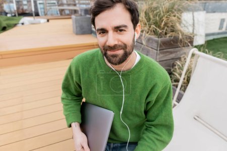 happy freelancer in earphones holding laptop on rooftop terrace in Vienna, Austria, remote work